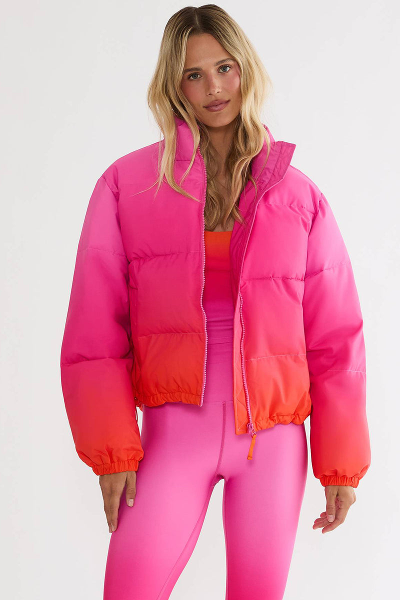 A woman modeling a cropped puffer jacket with a vibrant pink-to-orange gradient, paired with matching pink leggings, against a plain white studio background.