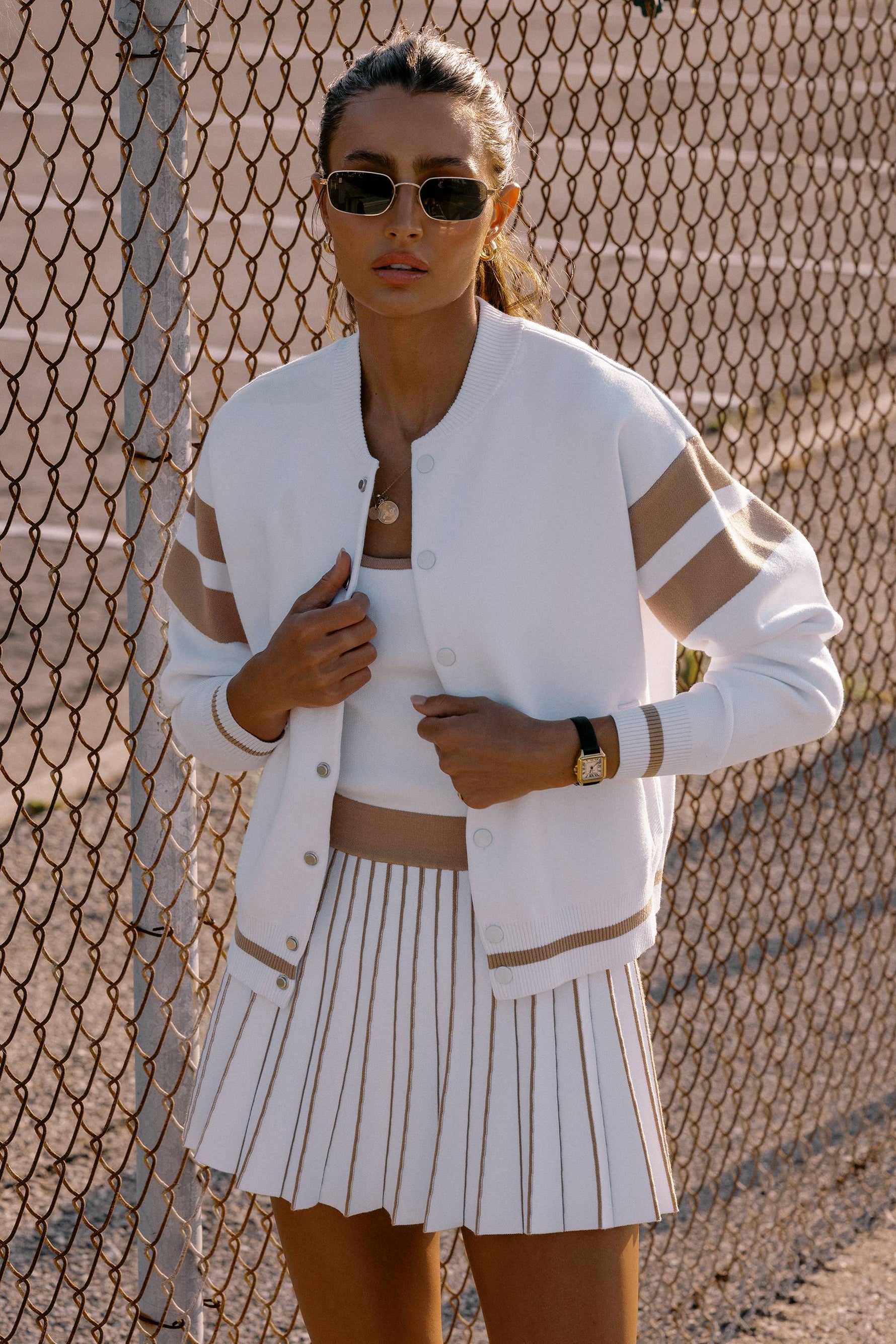 Woman wearing a white varsity jacket and pleated skirt against a chain-link fence.