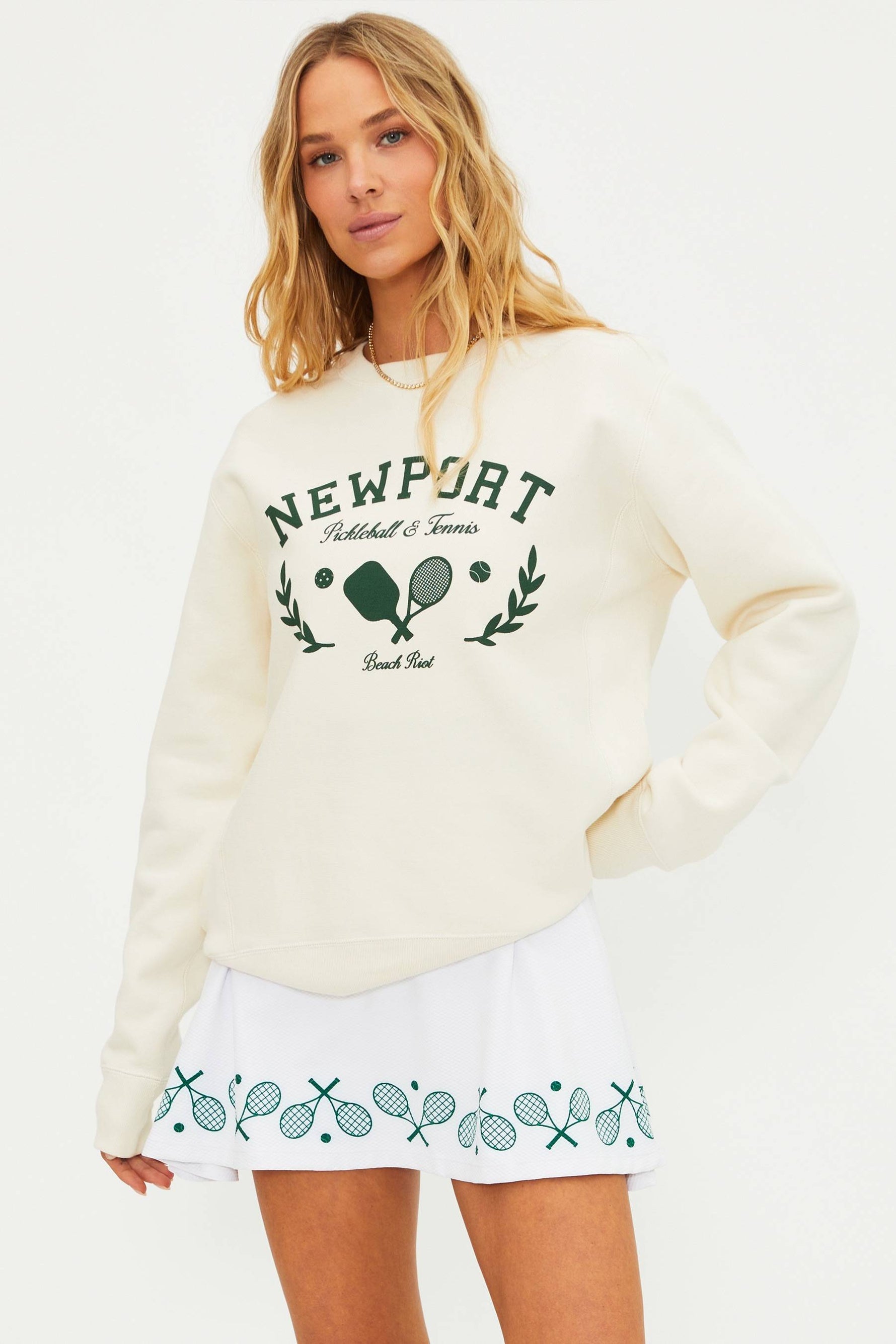 Woman wearing a cream sweatshirt with 'Newport' and tennis racket graphics, paired with a white skirt with tennis racket patterns.