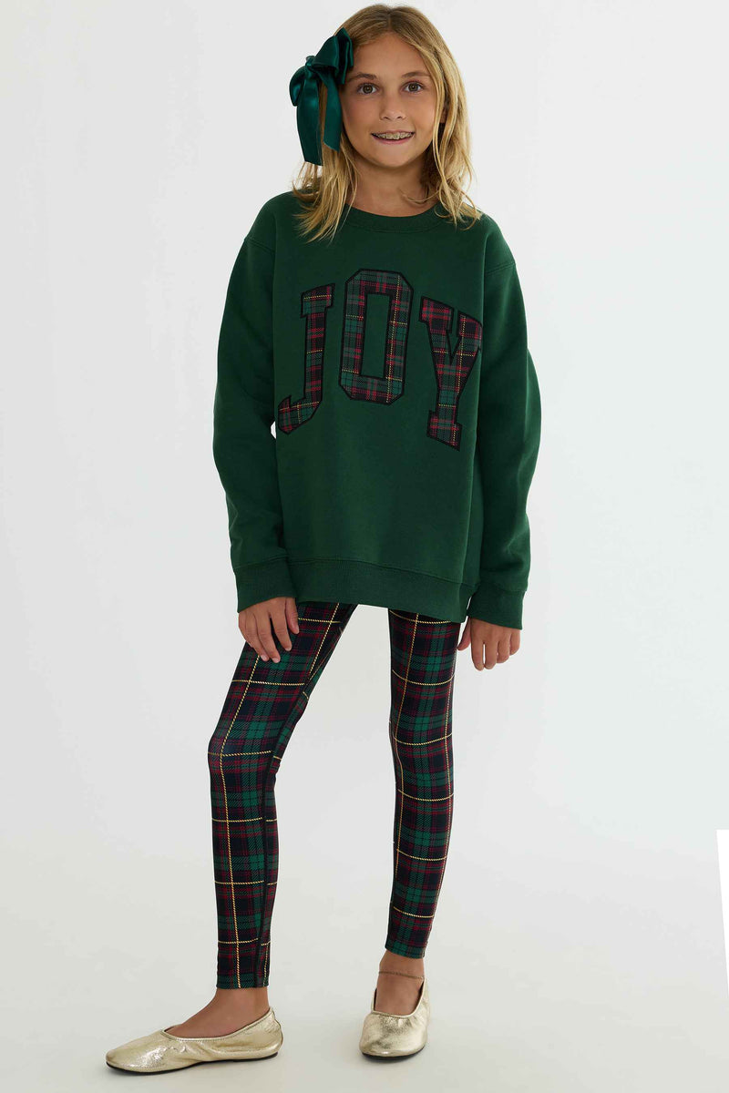 A young girl modeling a dark green crewneck sweatshirt with 'JOY' written in red and green plaid letters, paired with matching plaid leggings and gold metallic flats, against a plain white studio background.