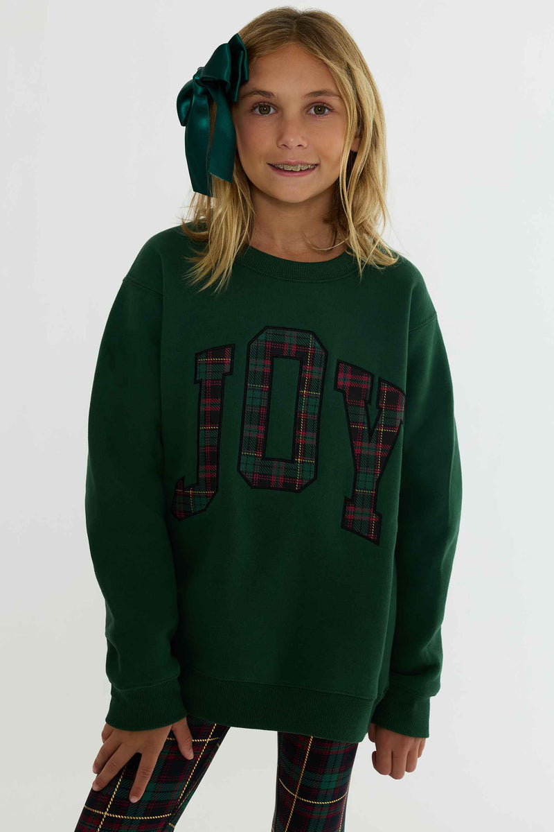 A young girl wearing a dark green crewneck sweatshirt with 'JOY' written across the chest in red and green plaid letters, paired with matching plaid leggings and a large green bow in her hair, against a plain white background.