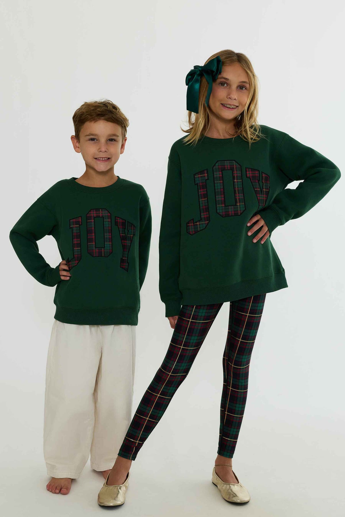 A boy and a girl modeling matching dark green holiday sweaters with 'JOY' written in red and green plaid letters, paired with plaid leggings and a matching bow headband.