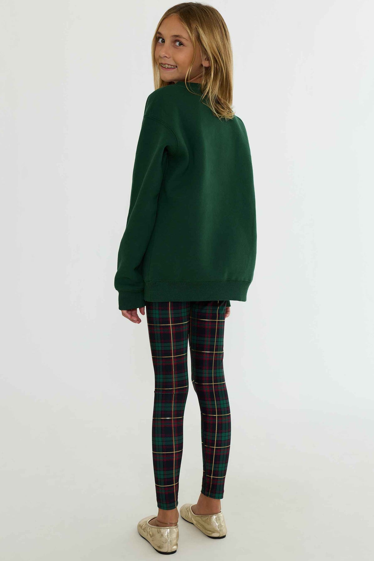 A young girl with blonde hair is shown from the back wearing an oversized dark green sweatshirt, green tartan plaid leggings, and metallic gold slip-on shoes, set against a plain white studio background.
