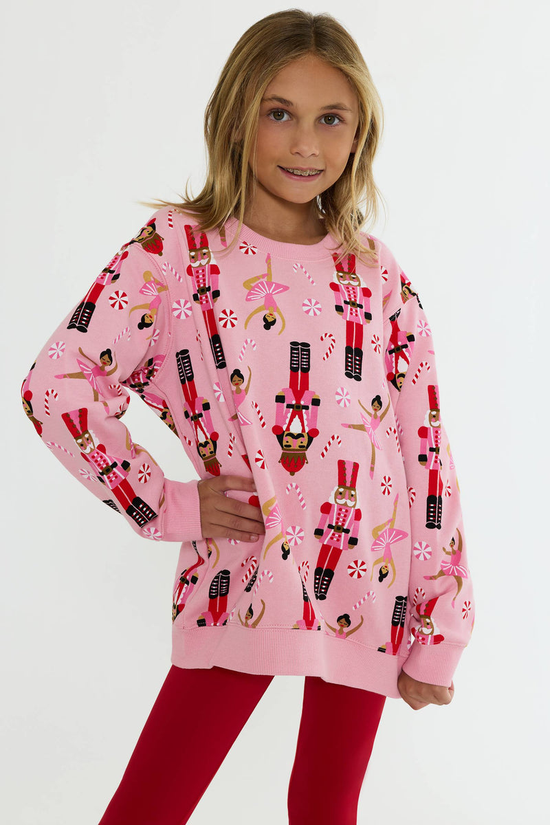 A young girl modeling a pink crewneck sweatshirt with a festive nutcracker and candy cane print, paired with red leggings, against a plain white studio background.