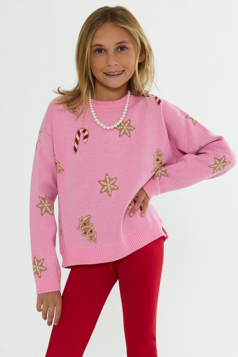 A young girl modeling a pink knit sweater with gold and red holiday motifs, paired with red leggings and a pearl necklace, against a plain white background.