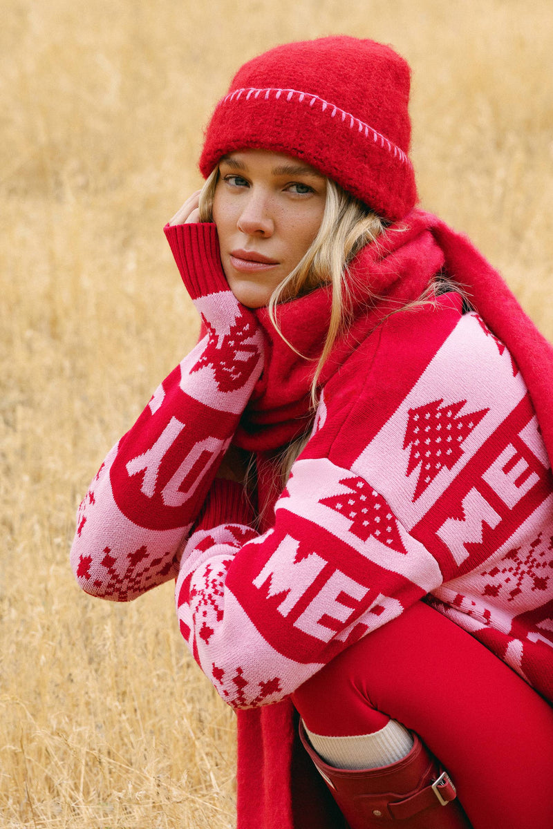 A woman wearing a festive red and white knit beanie, matching knit sweater with 'MERRY' and Christmas tree patterns, a red knit scarf, and red leggings, posed in a dry grass field.
