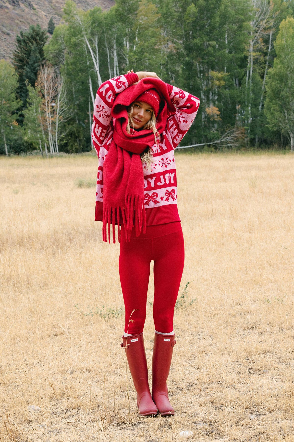 A woman stands in a dry grass field wearing a matching red knit holiday sweater, red scarf, red leggings, and red rain boots, with trees in the background.
