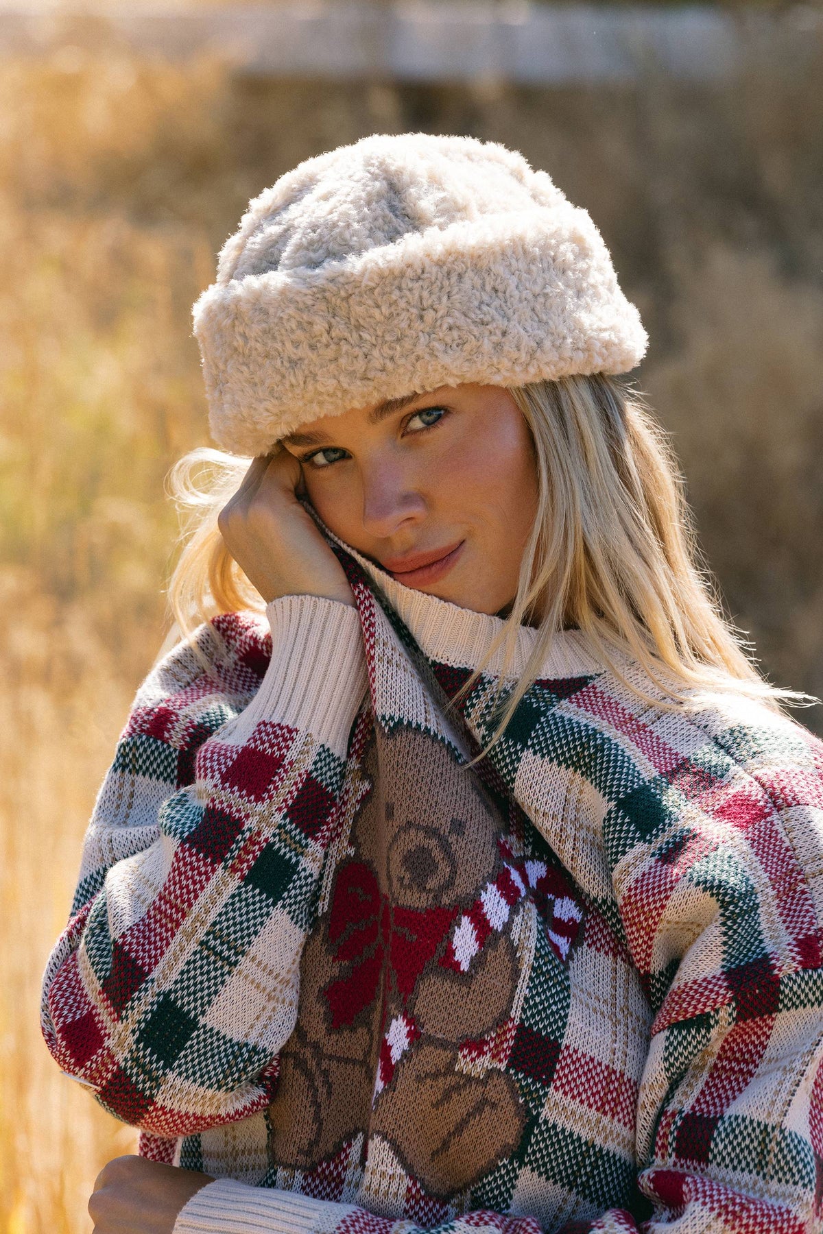 A woman outdoors wearing a chunky cream knit beanie and a red, green, and cream plaid sweater with a teddy bear graphic.