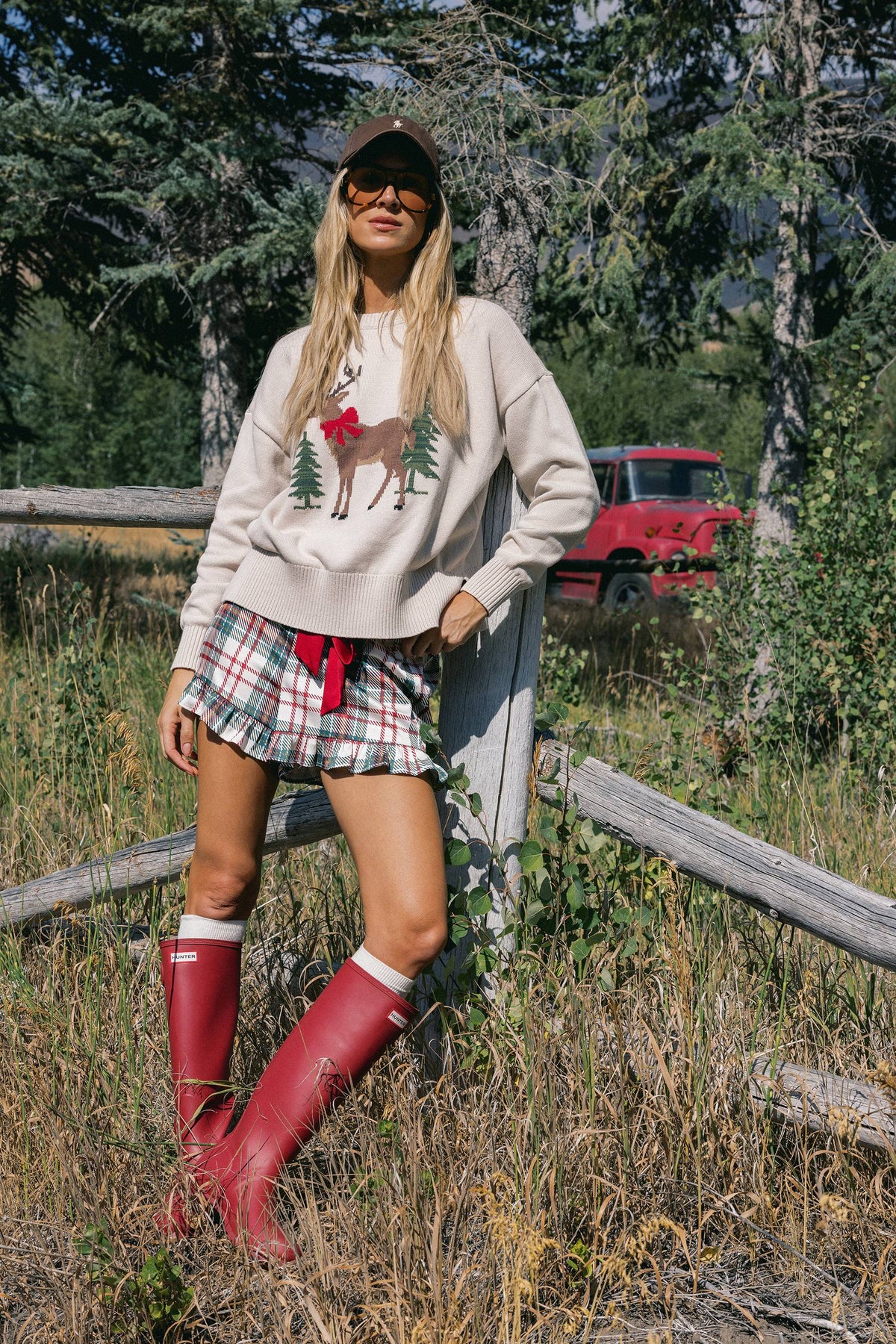 A woman stands outdoors in a grassy wooded area wearing a beige crewneck sweatshirt with a reindeer graphic, a red-and-white plaid mini skirt, and red rain boots. A red vintage truck is visible in the background behind trees.
