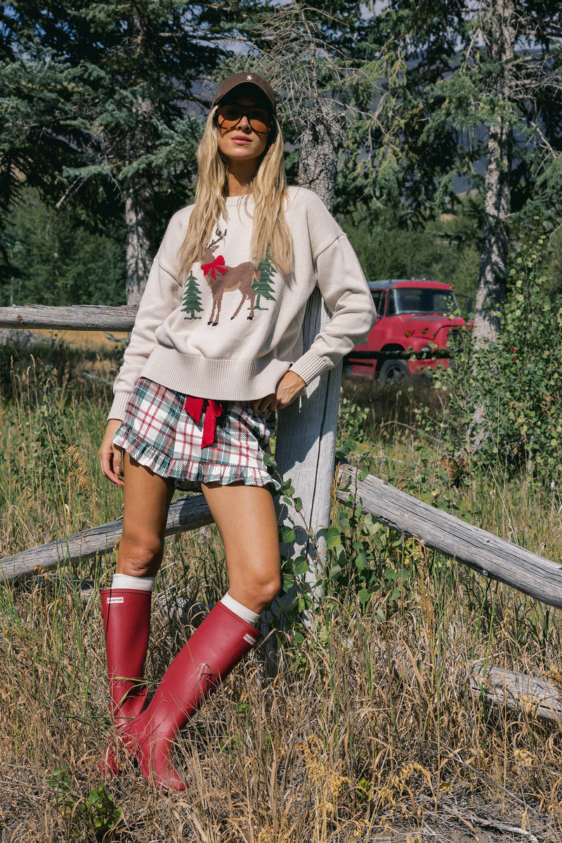 A woman stands outdoors in a grassy wooded area wearing a beige crewneck sweatshirt with a reindeer graphic, a red-and-white plaid mini skirt, and red rain boots. A red vintage truck is visible in the background behind trees.
