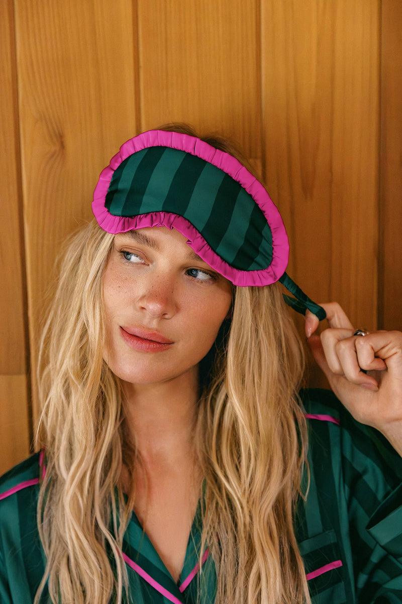 A woman with long blonde hair wearing a dark green and black striped sleep mask with a bright pink ruffled trim, posed against a warm wood-paneled background.