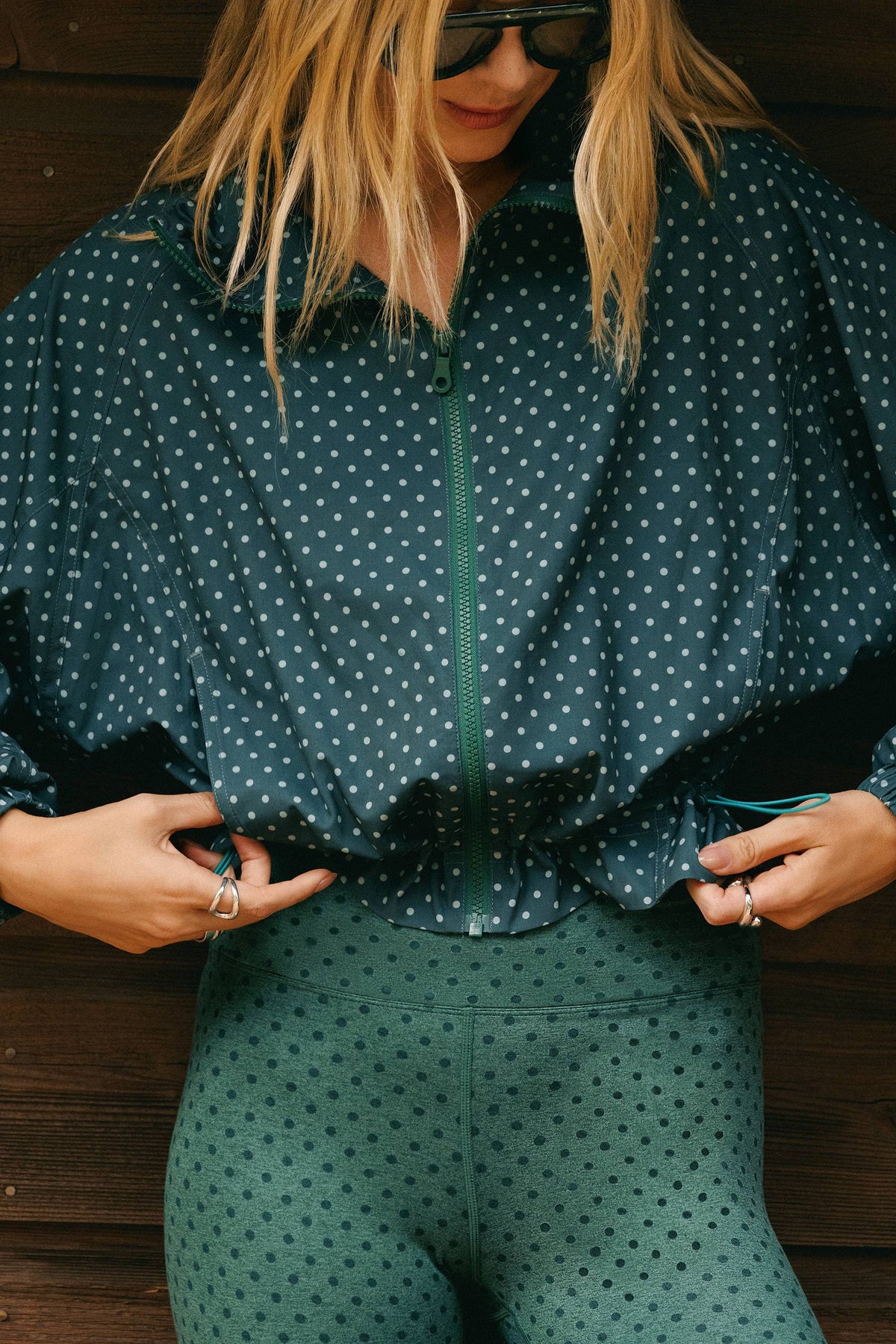 A woman wearing a dark teal green zip-up shirt and matching high-waisted leggings with a small white star pattern, posed against a dark wood plank background.