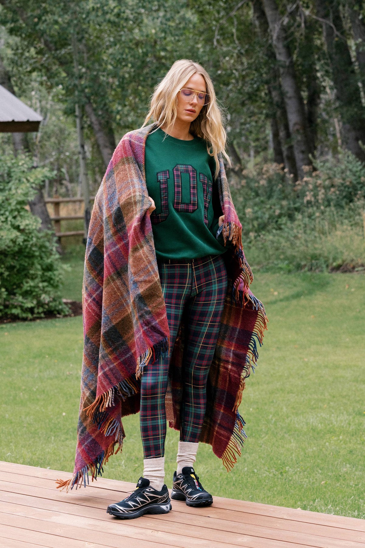 A woman stands outdoors on a wooden deck in a grassy park area, wearing a green crewneck sweatshirt with '107' in plaid, matching plaid pants, white socks, black sneakers, and holding a large multicolor plaid fringed blanket draped over her shoulders.