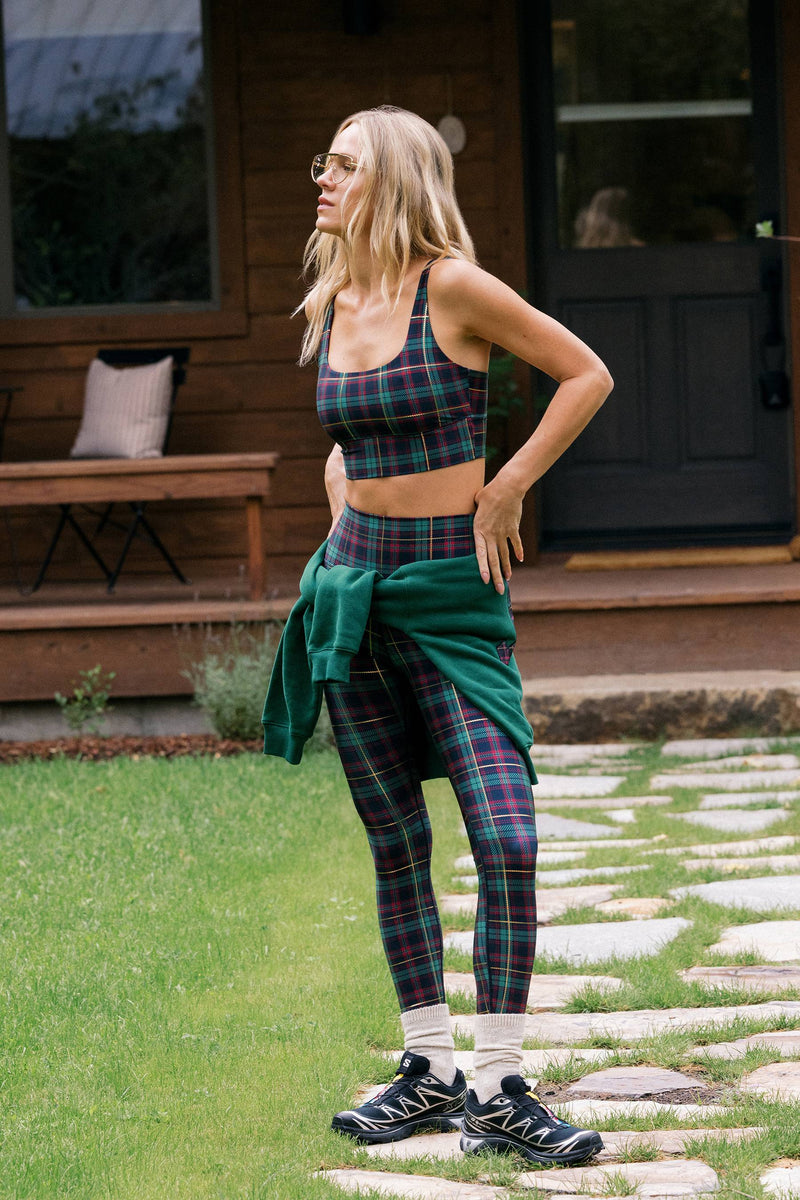 A woman stands outdoors in front of a wooden house, wearing a matching plaid sports bra and leggings set with a dark green sweater tied around her waist. The plaid pattern mixes green, red, and dark blue tones.