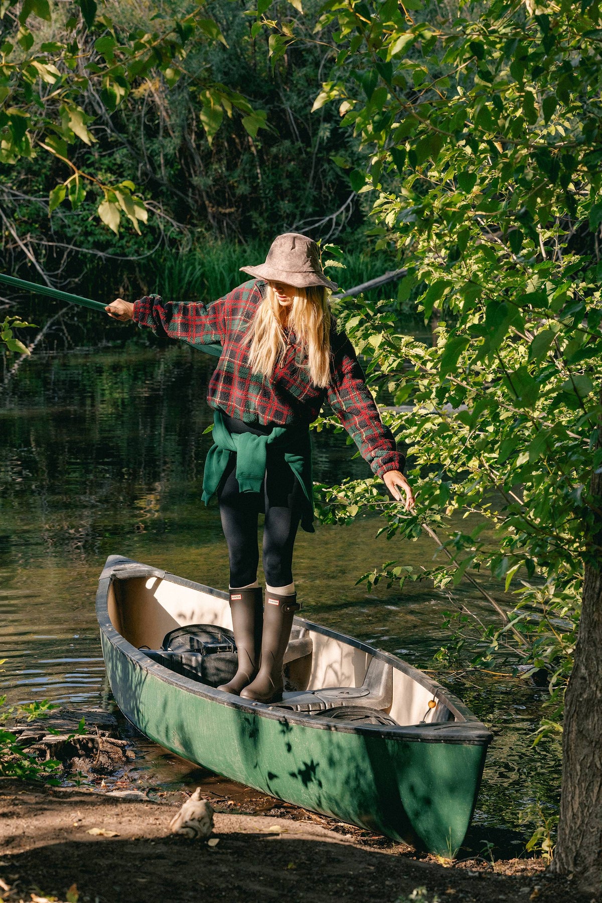 A woman stands in a green canoe on a river, wearing a red and black plaid flannel shirt, a brown wide-brim hat, a green belt, black leggings, and tall brown boots. Trees and foliage frame the scene on a sunny day.