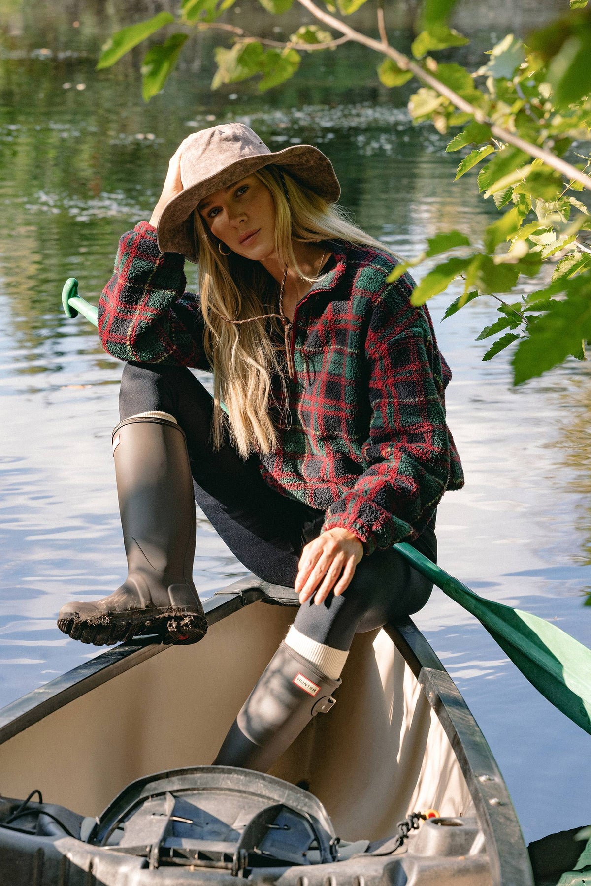 A woman sits in a canoe on a lake, wearing a red and green plaid flannel shirt, black leggings, tall dark rubber boots, and a wide-brimmed hat, with green leaves hanging into the foreground.