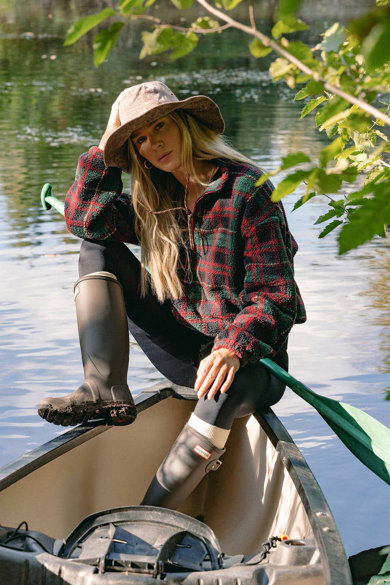 A woman sits in a canoe on a lake, wearing a red and green plaid flannel shirt, black leggings, tall dark rubber boots, and a wide-brimmed hat, with green leaves hanging into the foreground.