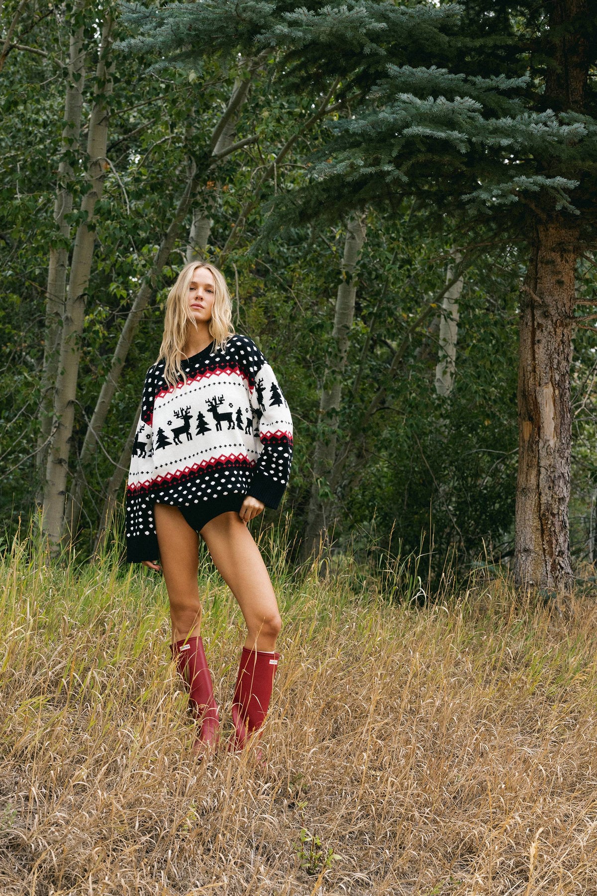 A woman stands in a grassy forest clearing wearing an oversized black, white, and red knit sweater with reindeer motifs, red knee-high socks, and no visible top underneath.