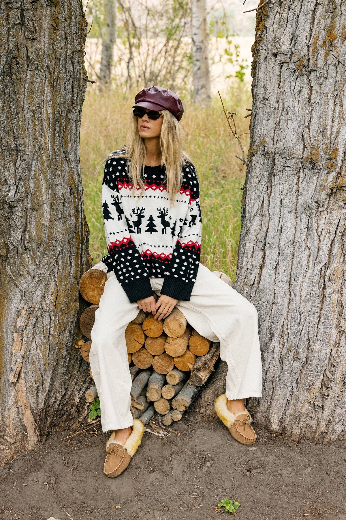 A woman sits on a stack of cut logs between two large tree trunks outdoors. She is wearing a black, white, and red patterned knit sweater with a burgundy beret, white pants, and tan slippers.