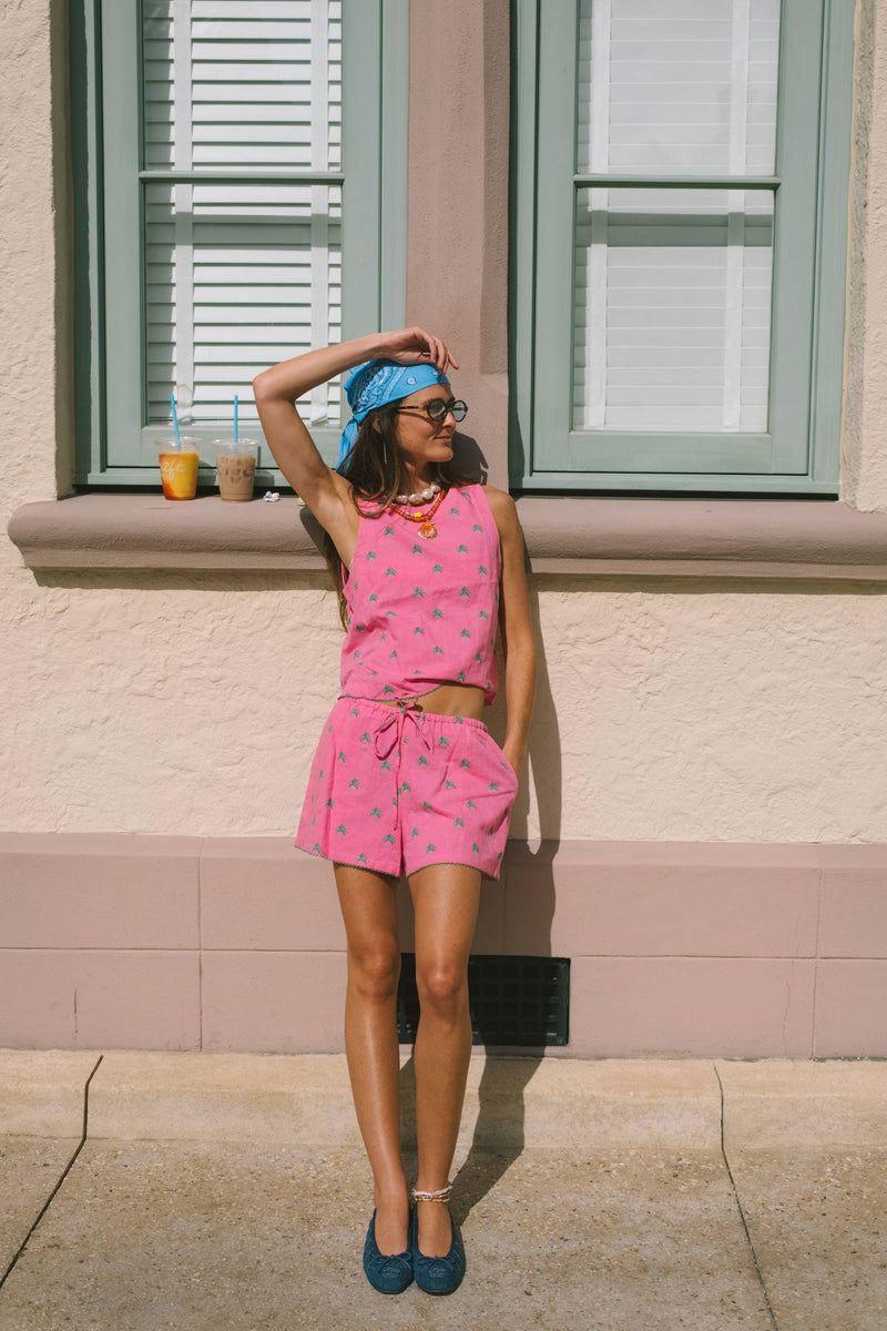 A woman stands outdoors against a beige stucco building wall with green shutters, wearing a bright pink two-piece outfit with a small repeating pattern, a blue headscarf, and blue shoes.