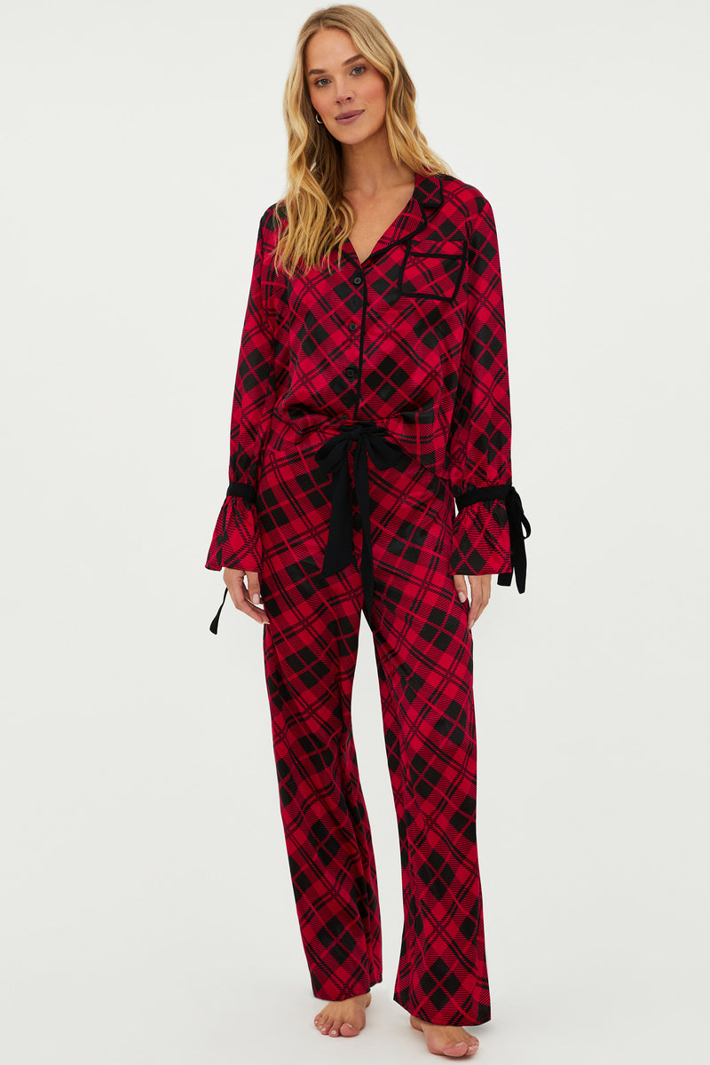 A woman standing, wearing a red and black plaid pajama set with a notched collar and long sleeves, featuring black velvet ribbon details at the cuffs and a black ribbon drawstring at the waist.