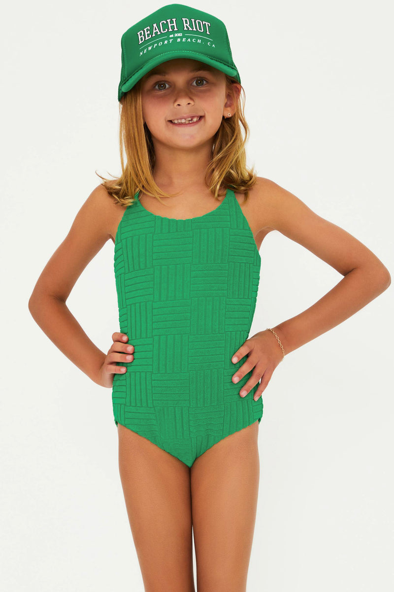 A child model wearing a green one-piece swimsuit with a textured pattern and crossed back straps. The model is also wearing a matching green cap with the text 'BEACH RIOT' on it.