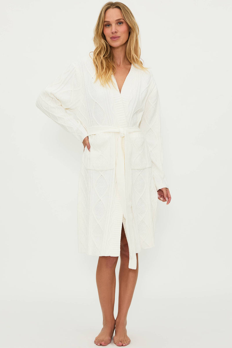 A female model wearing a white cream cable knit robe with a shawl collar and front pockets, belted at the waist.